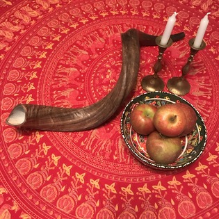 Shofar (ram's horn) candlesticks and bowl of three apples on a red tablecloth with round, manorla pattern