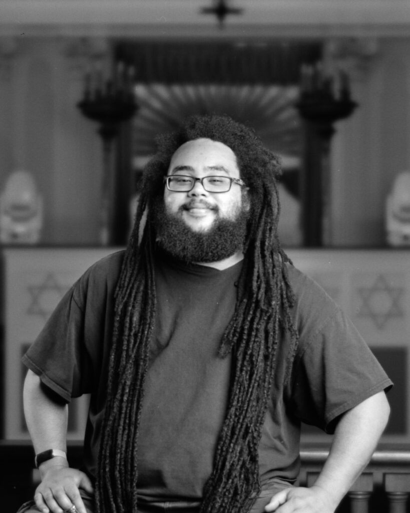 Black and white photo of a person with glasses a beard and dreadlocks looking at the camera