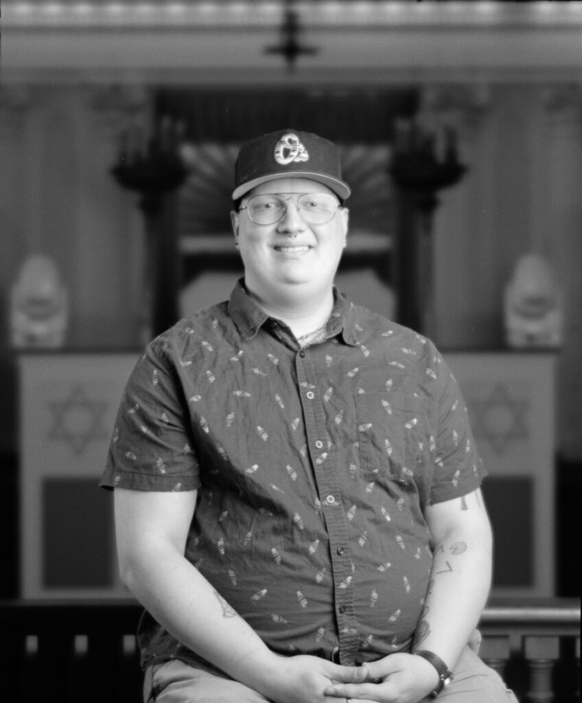 Black and white photo of a person in a button-up shirt and a baseball cap smiling at the camera