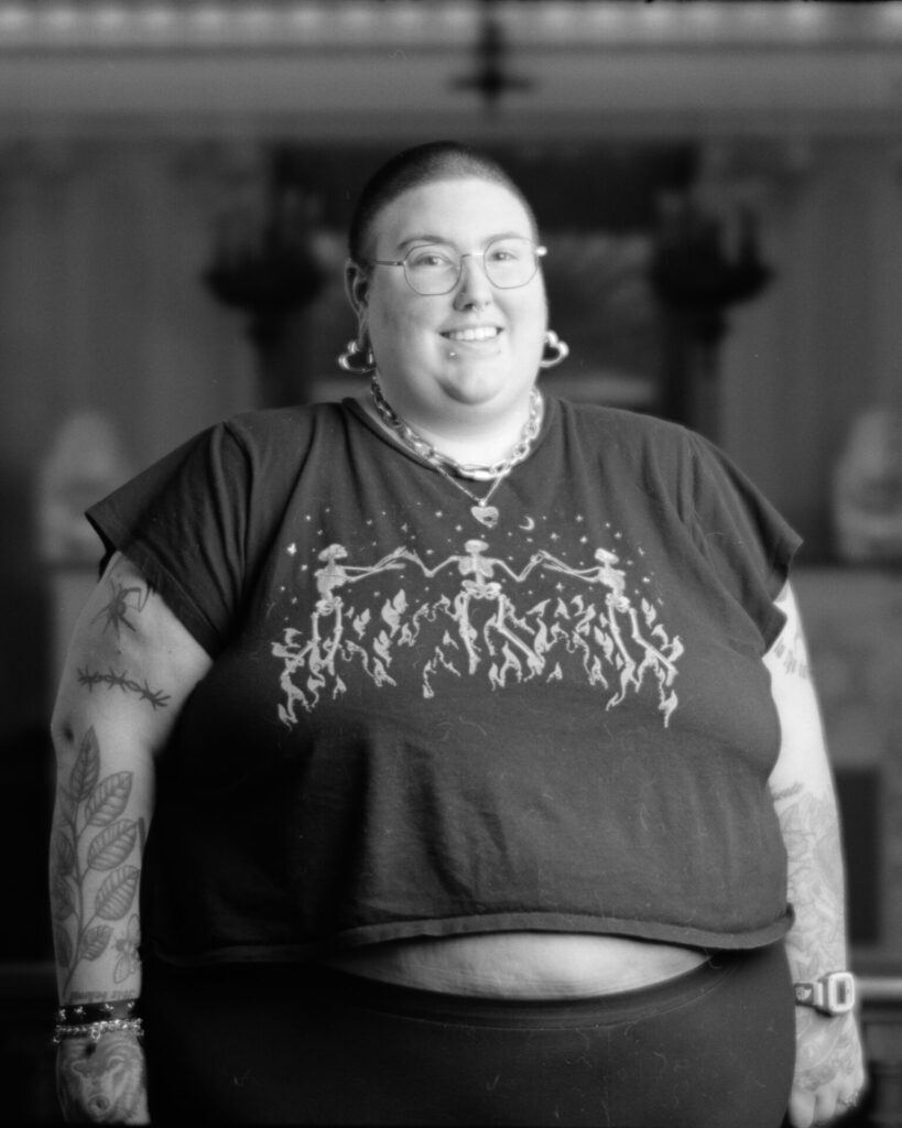 Black and white photo of a person in a dark tee shirt with tattoos on one arm smiling at the camera