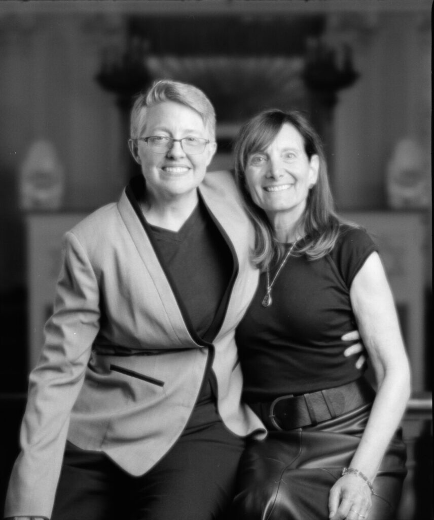 Black and white photo of two people smiling at the camera, one in a blazer, one in a dress