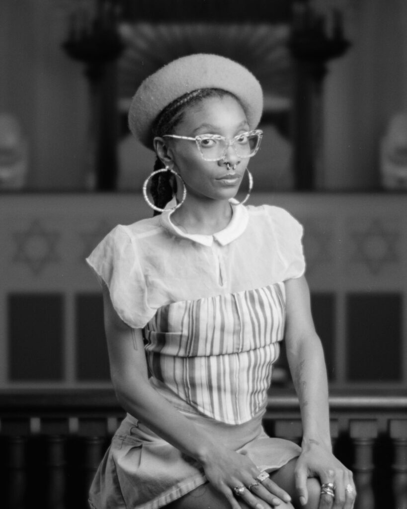 Black and white photo of a person in a hat and glasses smiling at the camera.