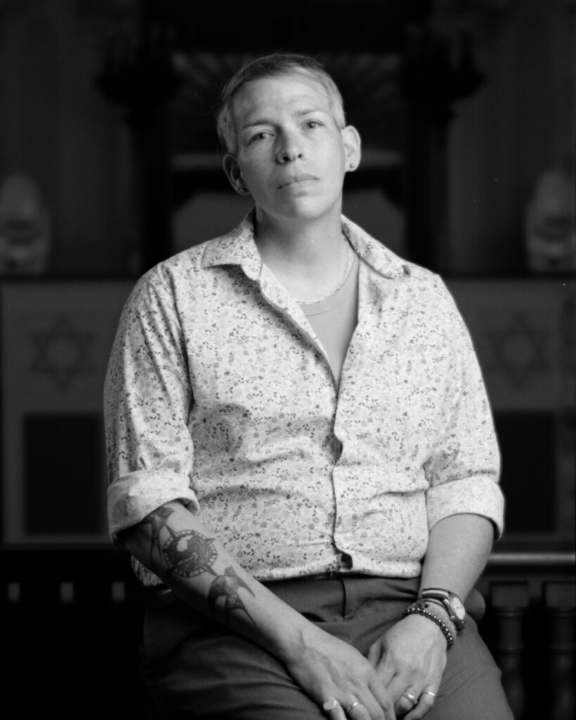 Black and white photo of a person in a button-up shirt with sleeves rolled to reveal tattoos on one arm looking at the camera