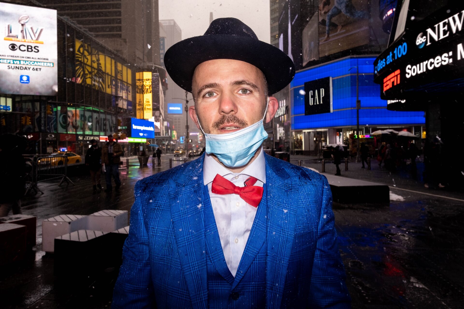 Rapper Kosha Dillz wearing a blue suit and red bowtie, with a mask pulled down around his chin
